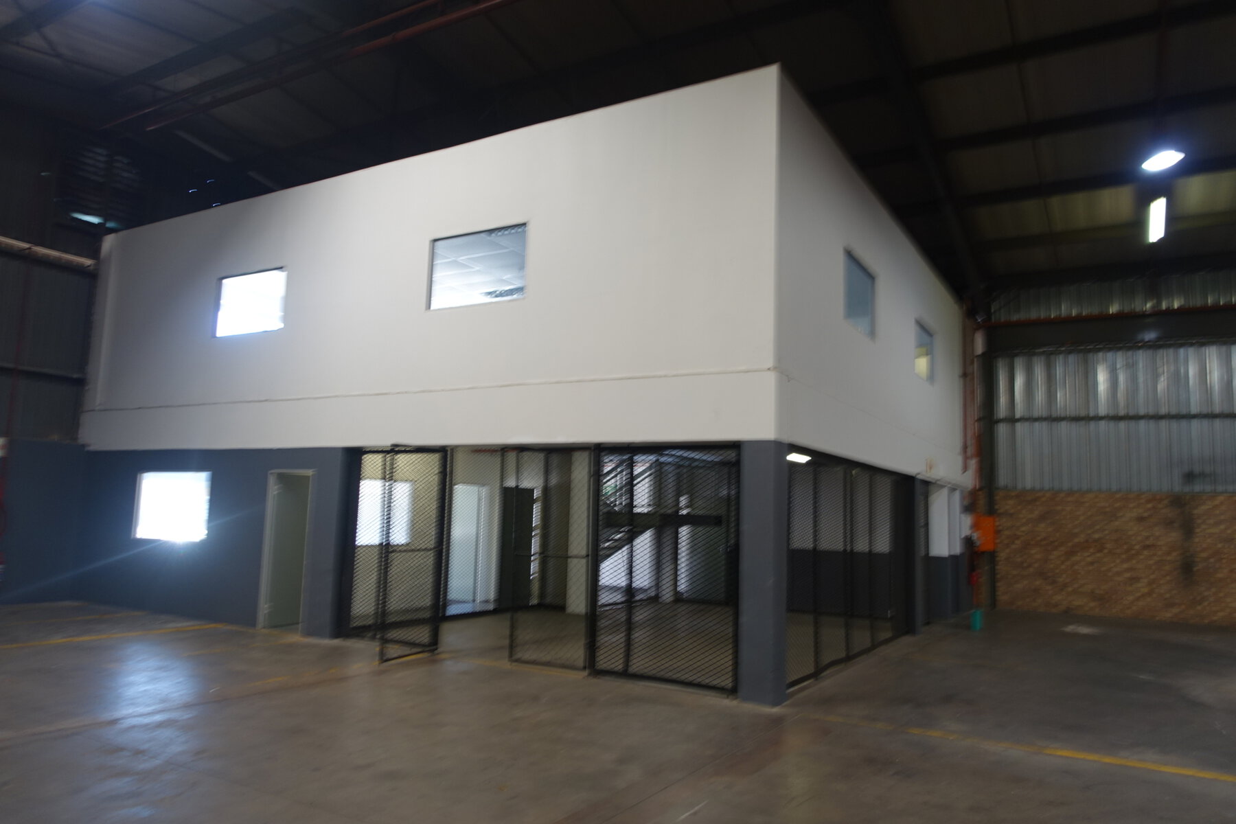 A contemporary office and warehouse building with large windows and glass doors, situated inside a spacious industrial warehouse.