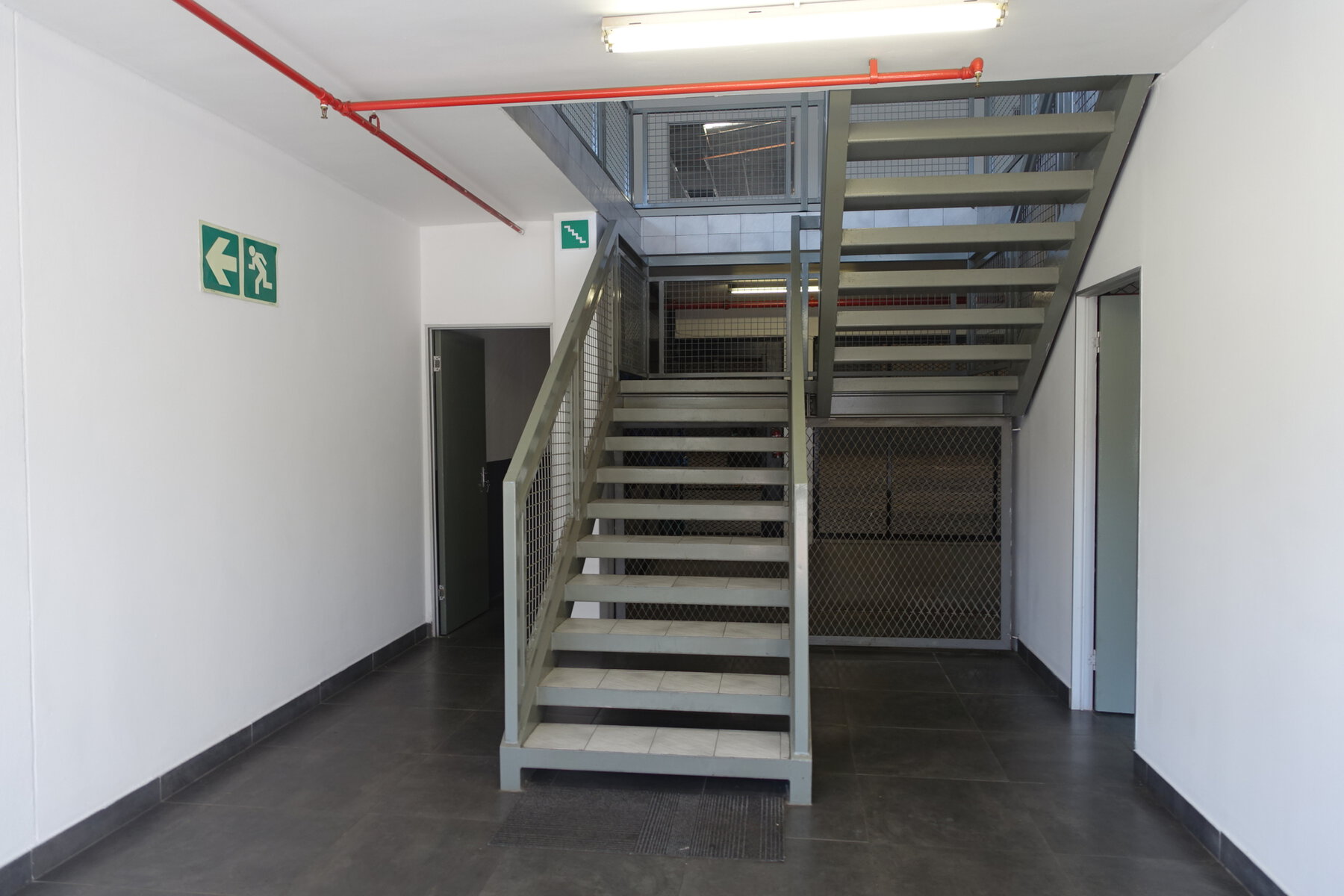 Steel staircase with metal mesh steps inside a contemporary office and warehouse space, featuring white walls, emergency exit signs, and a clean, organized environment.