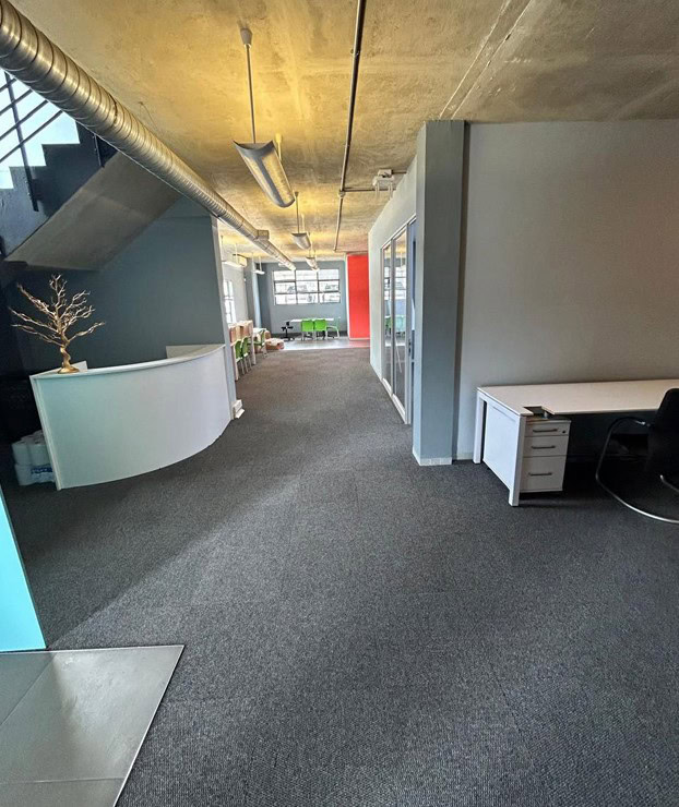 Modern industrial office space with open layout and large windows. Office interior with contemporary design, gray walls, and carpeted flooring.