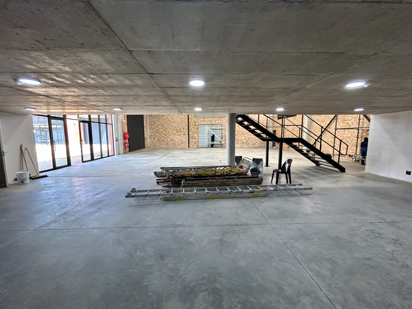 Spacious industrial space featuring unfinished concrete floors, ceiling lighting, and a staircase leading to an upper level, indicating ongoing development.