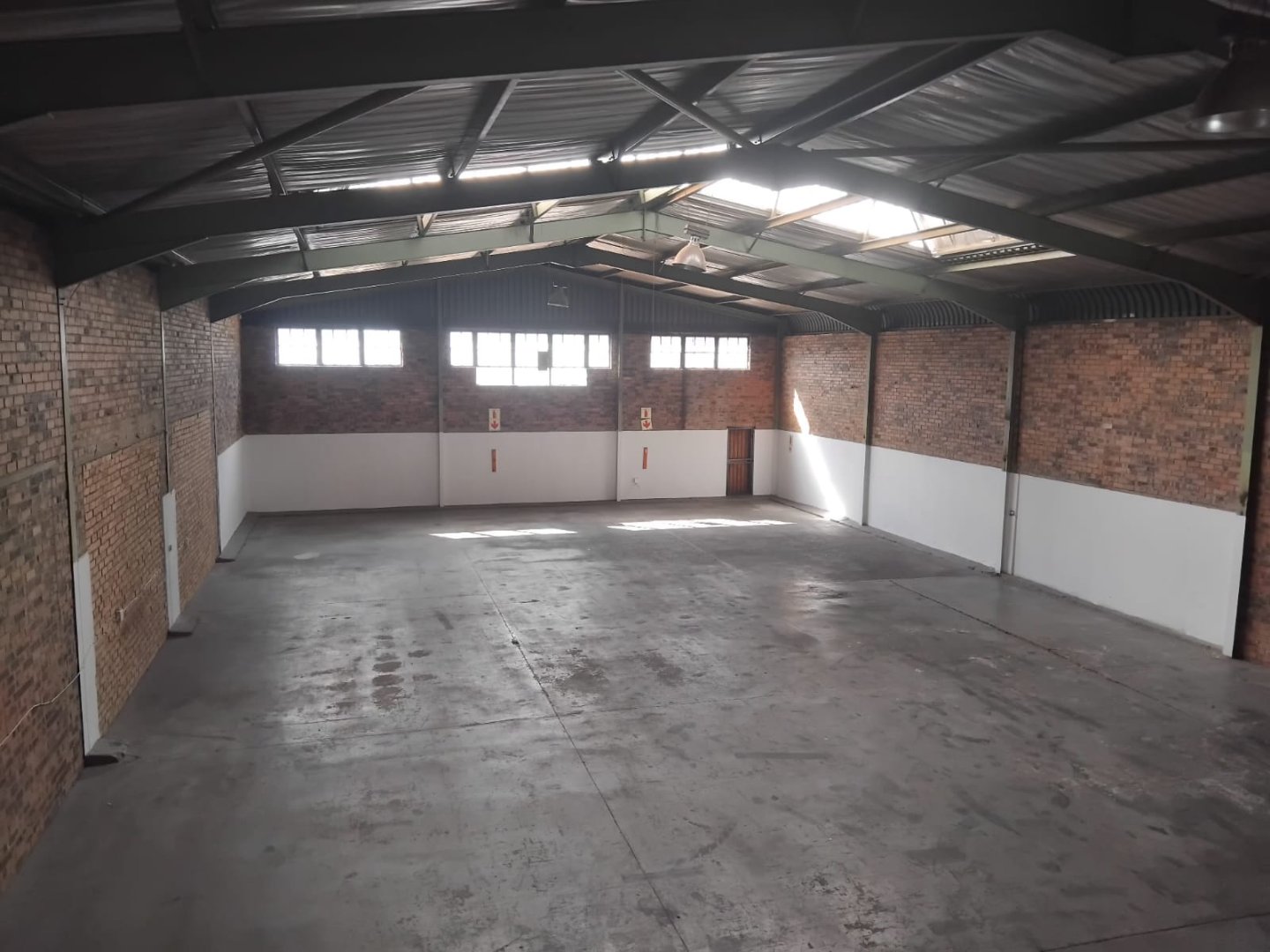Spacious industrial warehouse interior with brick walls and metal roof A large, empty warehouse space featuring brick walls, a high metal roof with skylights, and a concrete floor, ideal for storage or industrial use.