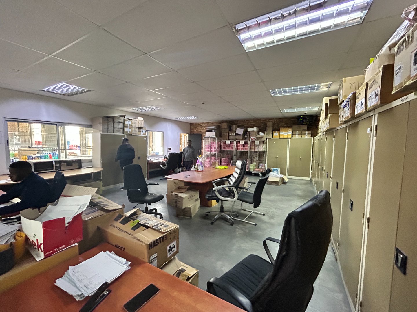 Office workspace with desks, chairs, and storage cabinets in a busy warehouse environment. An organized office area within a warehouse, featuring desks, chairs, storage cabinets, and boxes, illustrating a functional workspace for administrative tasks.