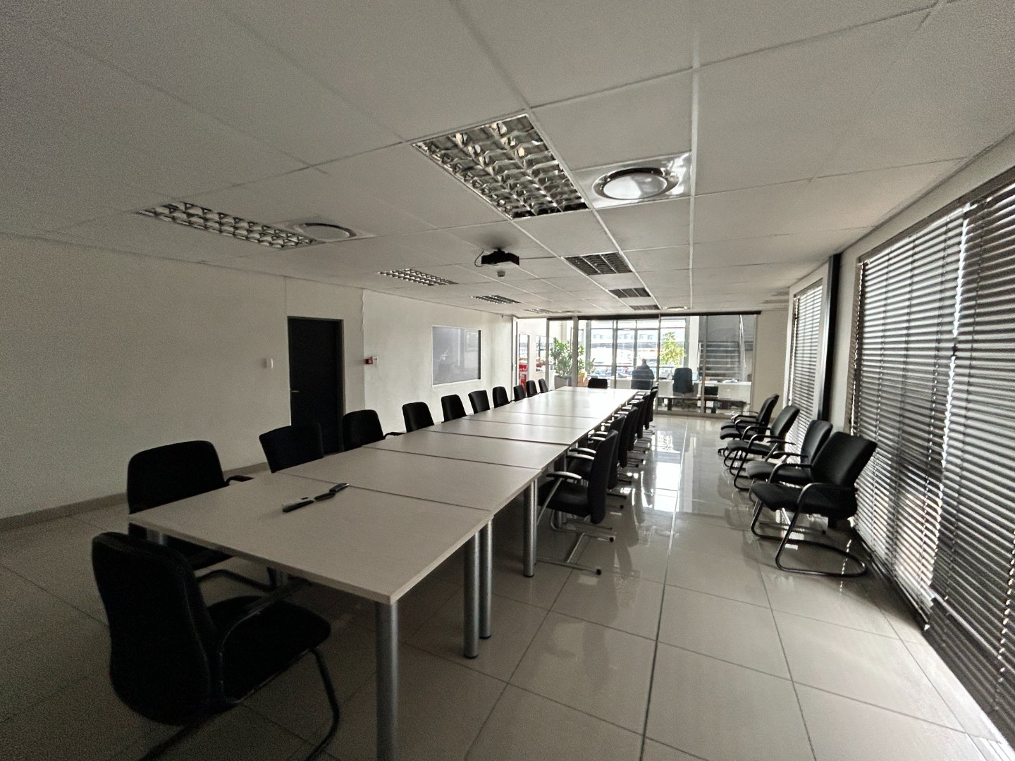 Modern office conference room with large table and black chairs, bright natural light, and window blinds. Office conference room with a long table, black chairs, and ample natural light from large windows, ideal for meetings and collaborative work.