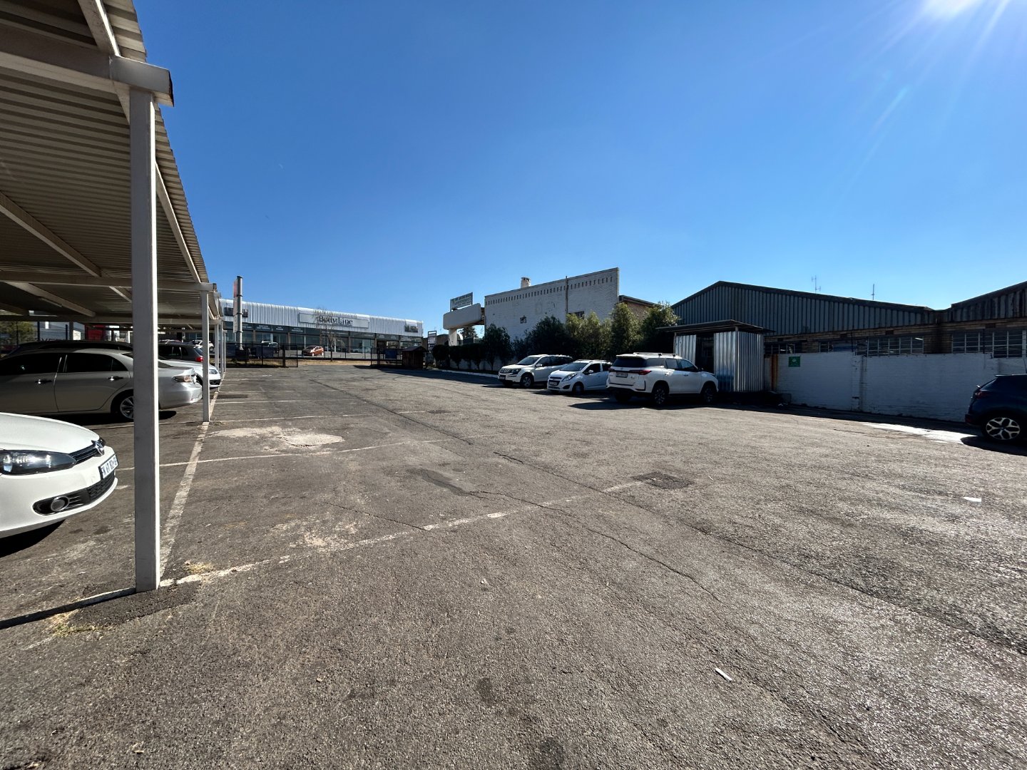 Spacious outdoor parking lot with multiple cars and industrial buildings in the background under a clear blue sky.