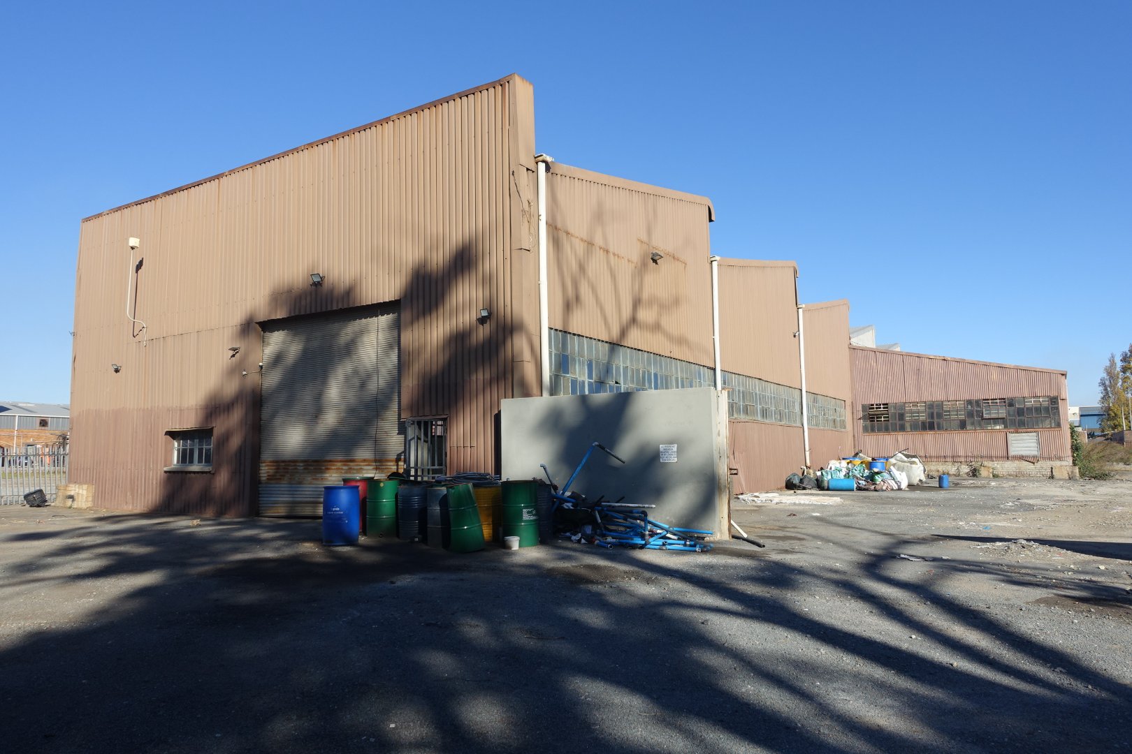 Exterior view of an industrial warehouse with large beige metal walls and a loading dock, surrounded by barrels and equipment, under a clear blue sky.