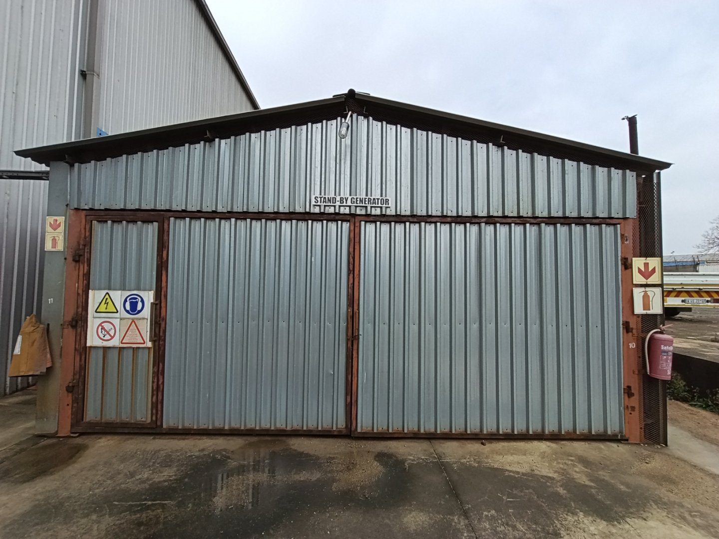 Large corrugated metal storage shed at Currie Group with safety signs and fire extinguisher, used for warehouse and office space since 1887.