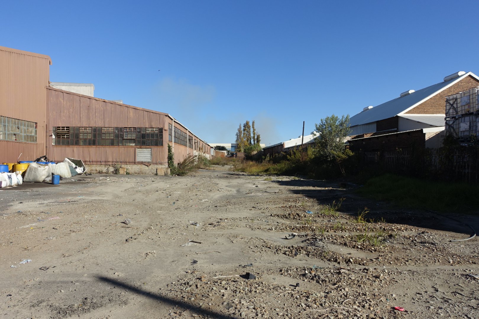 Open land between industrial warehouses and residential buildings under clear blue sky.