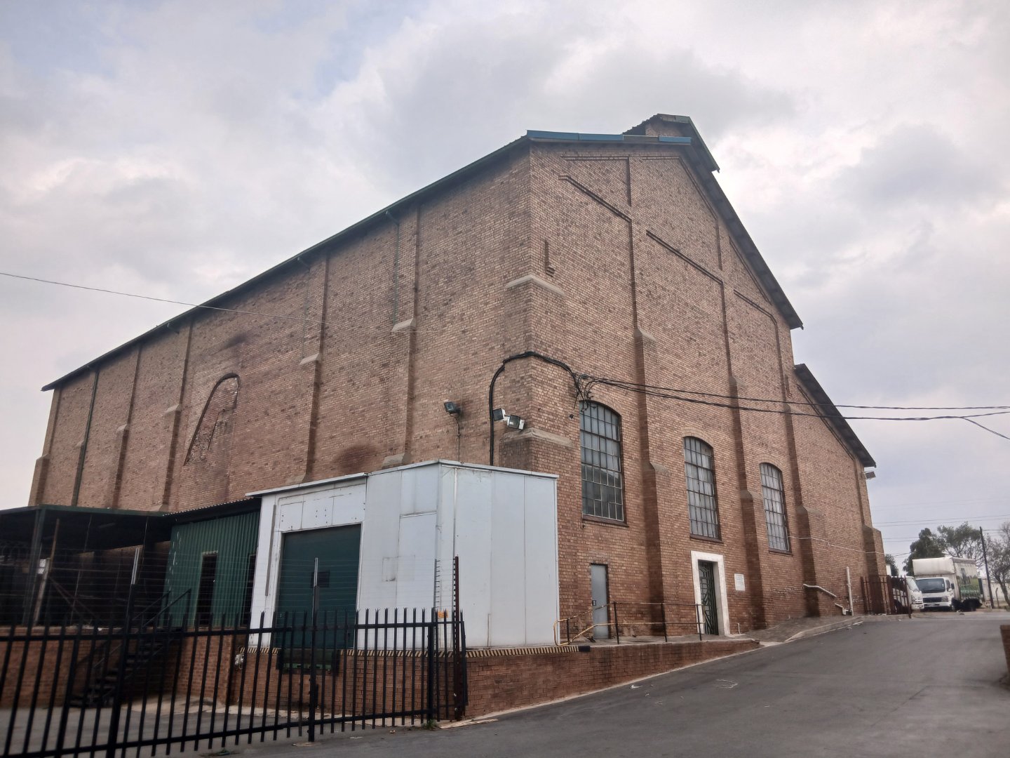 Large historic brick warehouse with multiple windows and a small white extension, located in an industrial area with trucks in the background.