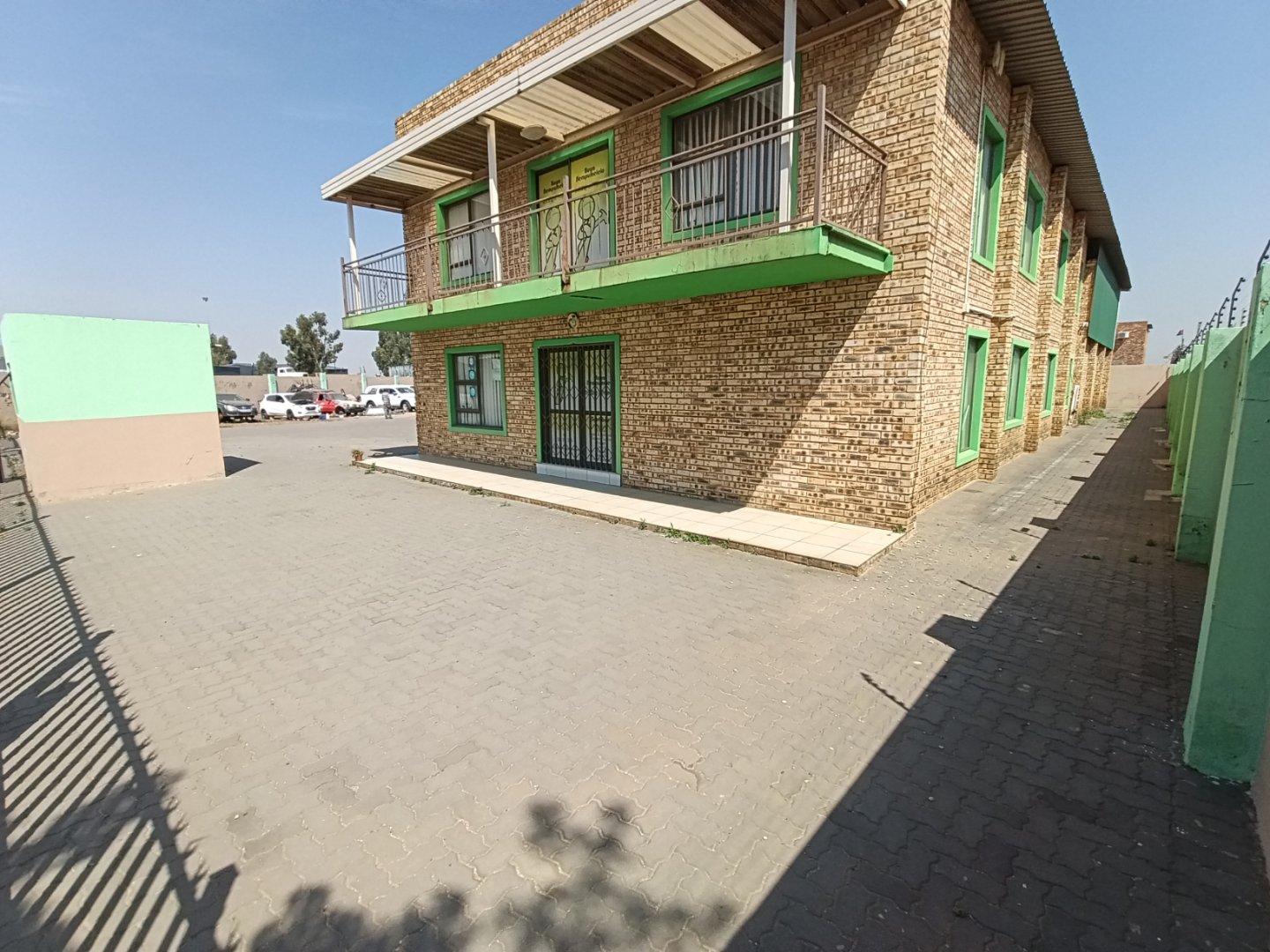 Two-story brick building with green accents and balconies, located in Alrode, South Africa, with a paved parking area and surrounding fencing.
