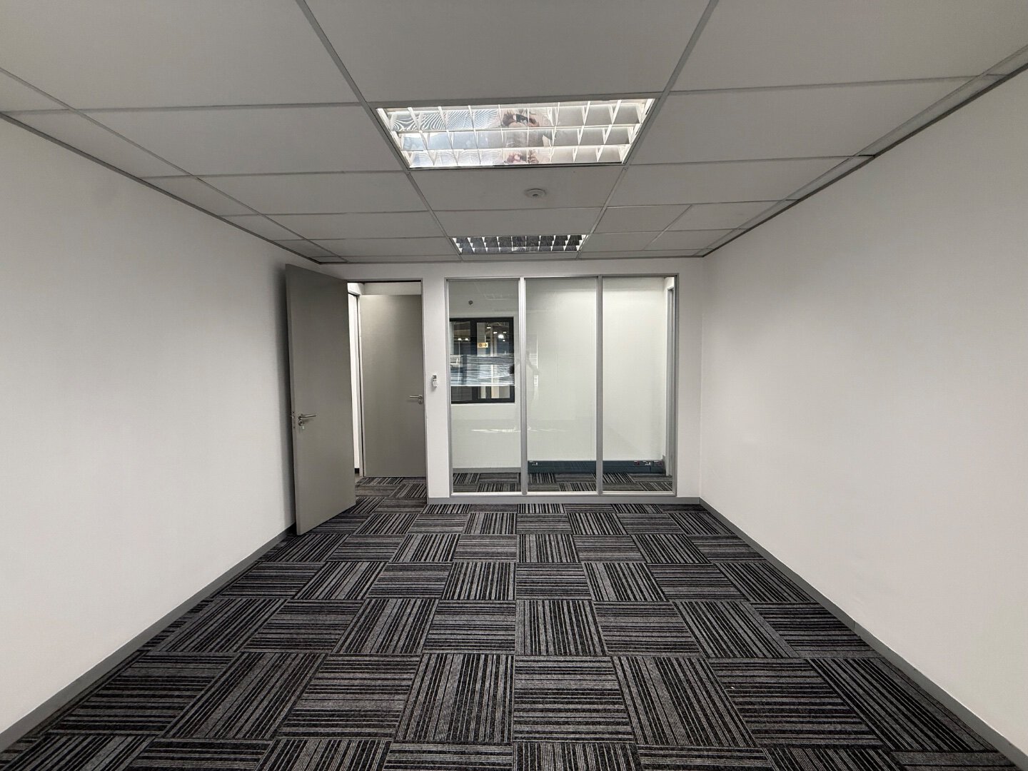 Modern Office Interior with Glass Doors Spacious office room with white walls, black patterned carpet, and glass doors leading to another office space, ideal for corporate environments.