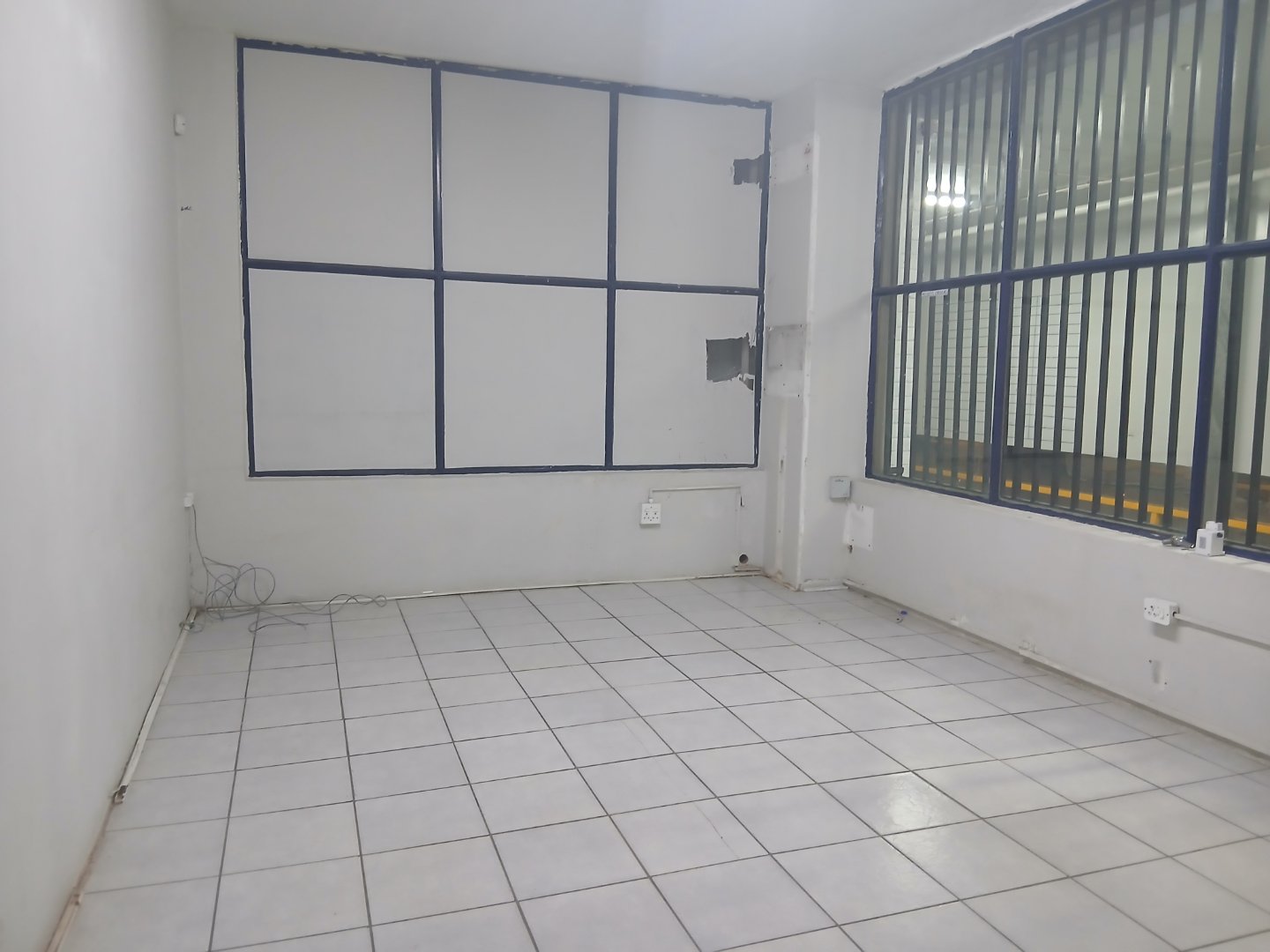 Empty office room with white tiled floor, large window with black bars, and a white wall with a window frame.