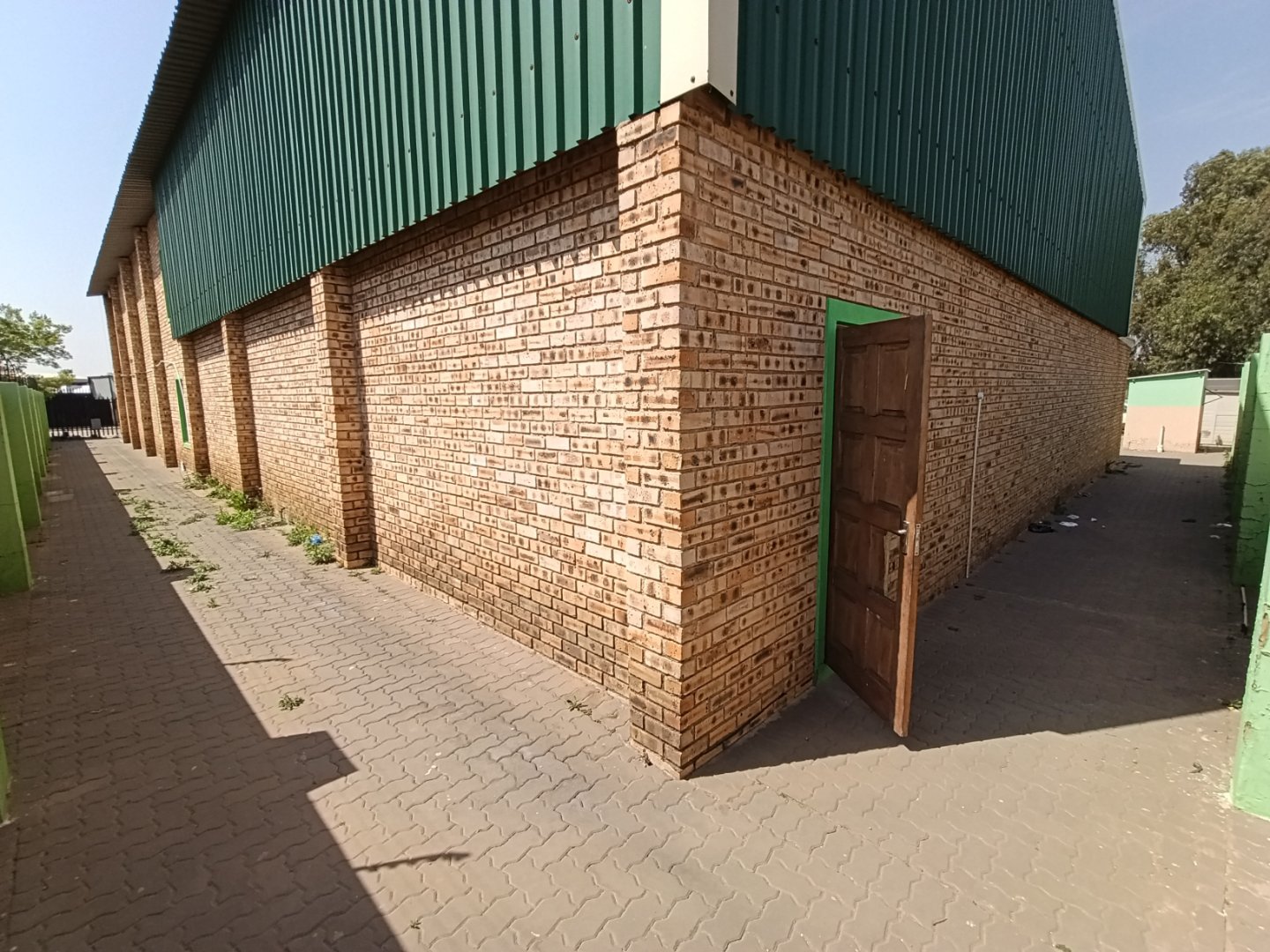 Exterior view of a brick warehouse with a green metal roof and a partially open wooden door, located in an industrial area.