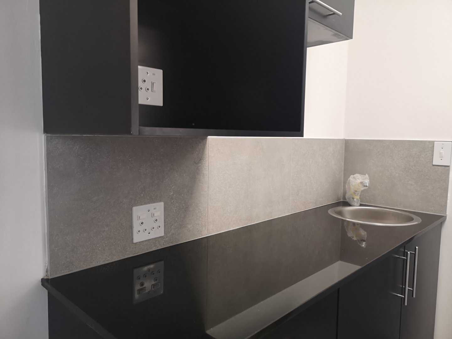 Modern Office Kitchen Area Contemporary office kitchen with black countertop, grey backsplash, and stainless steel sink, featuring electrical outlets and cabinetry.