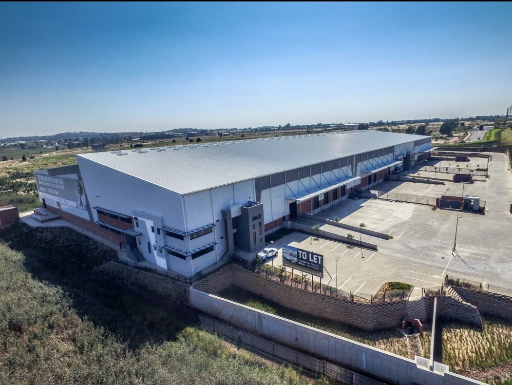 A large, modern warehouse with a white roof and multiple loading bays, situated in an industrial area with ample parking and clear skies.