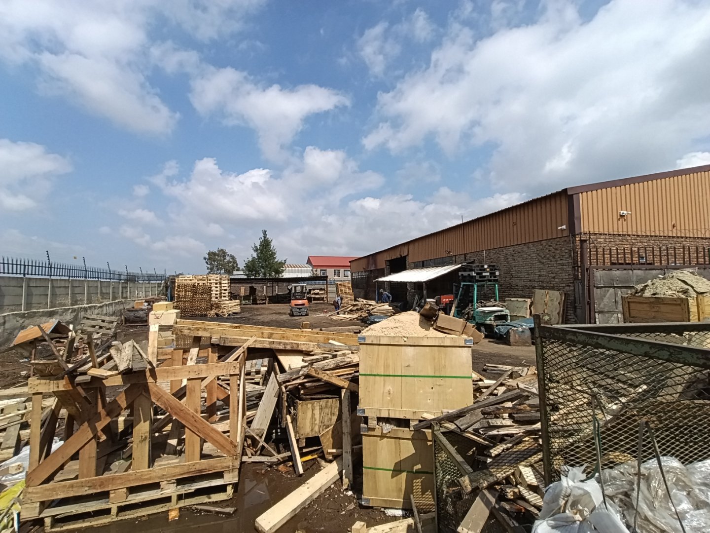 Construction site of an industrial warehouse with building materials, machinery, and partially built structures under a partly cloudy sky.