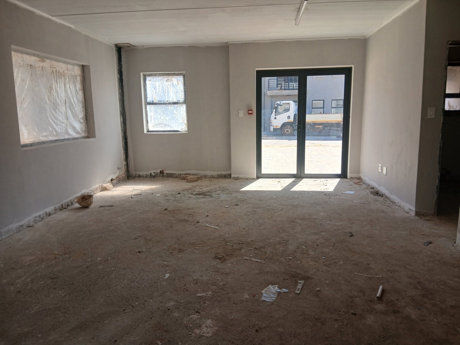 Interior view of a modern office and warehouse under construction with unfinished floors and walls, large windows, and a glass door leading outside.