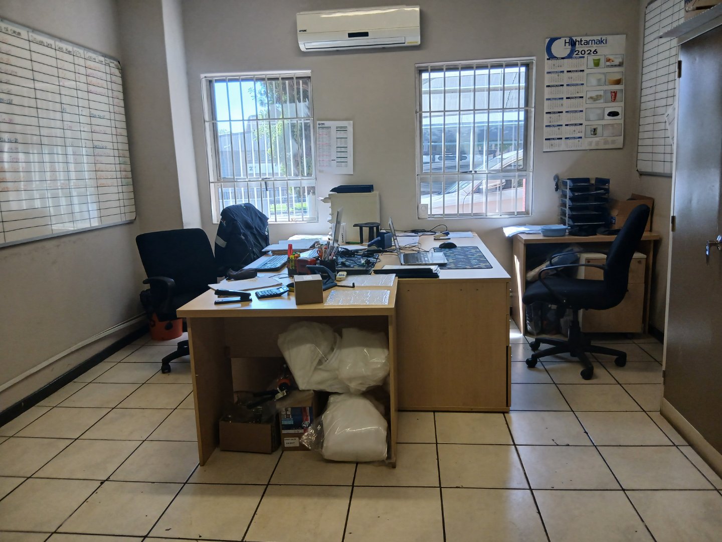 Interior view of a small office with two desks, chairs, and windows showing an outdoor view, featuring typical office equipment and supplies.